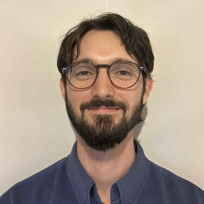 Dr Toby Muia smiles at the camera. He has dark short hair, a moustache and beard, and is wearing glasses